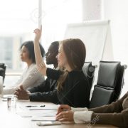 Smiling curious young businesswoman raising hand at multiracial group meeting engaging in offered activity, voting as volunteer or asking question at corporate business training, seminar or workshop